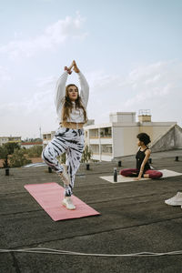 Full length of female friends practicing tree pose while friend exercising on rooftop