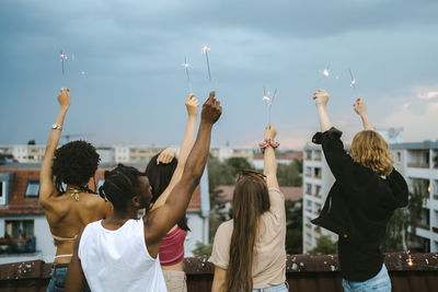 Rear view of multiracial friends with holding lit sparklers hands raised on rooftop