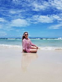 Full length of woman wearing sunglasses sitting at beach against sky