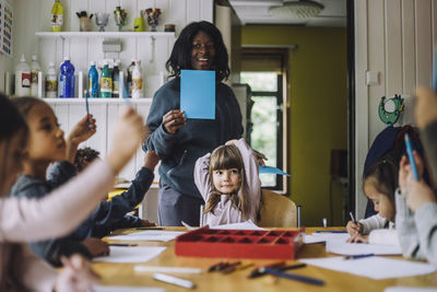 Happy teacher showing colored paper to students while teaching them in day care center