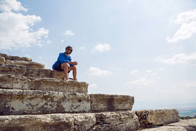 Man in a blue linen shirt and shorts sits on an old stone staircase of the colosseum in pamukkale