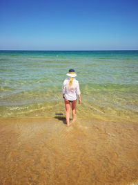 Rear view of young woman walking at beach against clear blue sky during sunny day