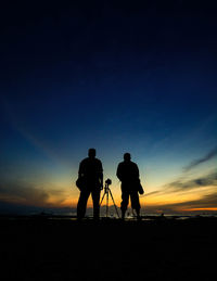 Full length of silhouette male friends standing amidst tripod at beach during sunset