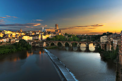 Bridge over river by buildings against sky at sunset