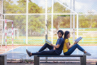 Young couple sitting on bench against fence