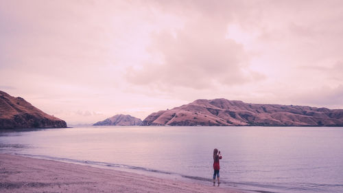 Woman standing by lake against cloudy sky during sunset