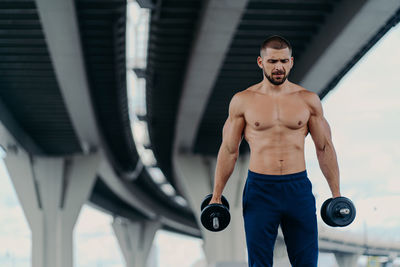 Shirtless young man lifting dumbbells while standing outdoors