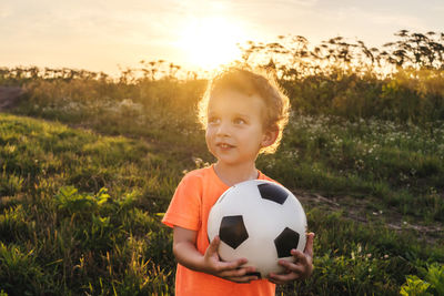 Cute boy holding ball while standing on grassy land at sunset