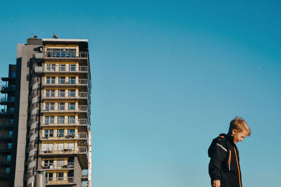 Low angle view of boy standing by building against clear blue sky