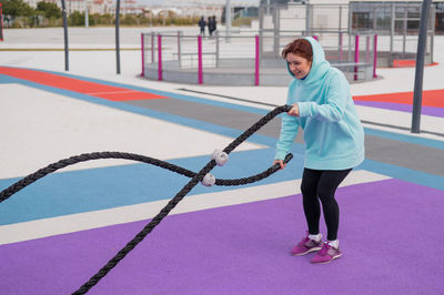 Caucasian woman in a mint sweatshirt is training with battle ropes at the sports ground. 