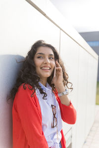 Smiling young woman talking on phone standing by wall outdoors