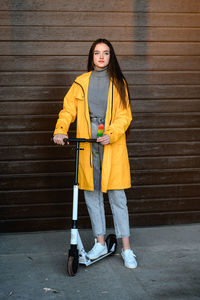 A girl in a yellow raincoat stands on a white scooter and holds colorful ice cream in her hands