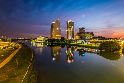 River by illuminated buildings against sky during sunset