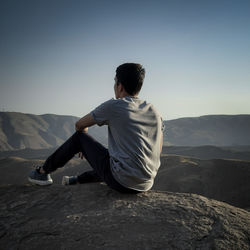 Rear view of man sitting on mountain against clear sky