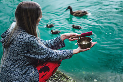 High angle view of young woman holding bowl while crouching by lake