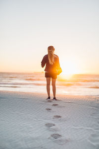 Rear view of young woman with skateboard standing at beach against clear sky during sunset
