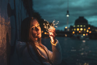 Portrait of young woman holding illuminated string light at night