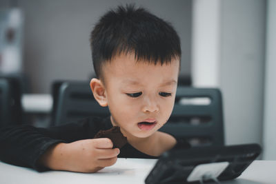 Cute boy eating biscuit while watching smart phone at home