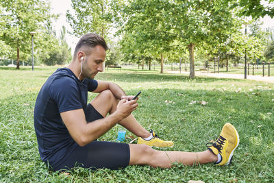 Man with his smart phone sitting on the grass in a park after training.