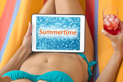 Midsection of young woman wearing bikini using digital tablet while resting on towel outdoors