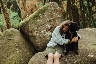 Girl hugging a black dog while sitting on top of a rock in the forest