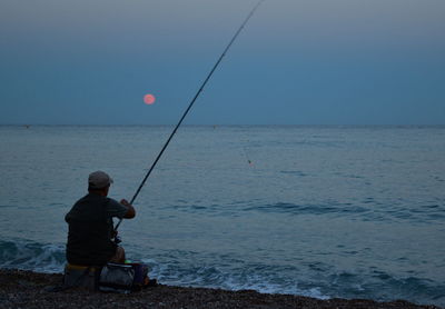 Rear view of man fishing in sea against clear sky
