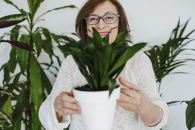 Smiling senior woman holding plant against wall at home