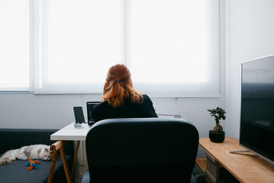 Back view of young female entrepreneur working on netbook at desk with tablet and smartphone at home