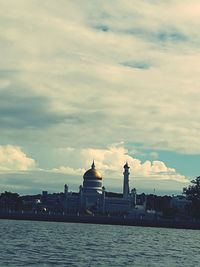 View of buildings by river against cloudy sky