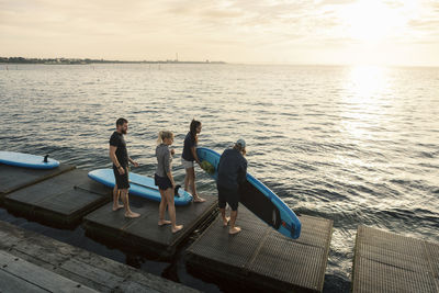 Male and female friends carrying paddleboard while standing on pier by sea