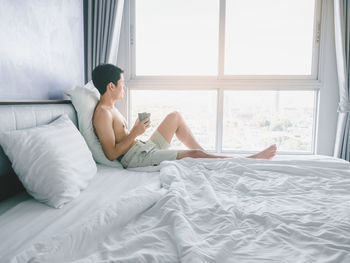 Asian young man with tattoo holding coffee cup on the bed looking out window in morning with sunrise