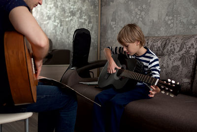 Young male musician teaches little boy student how to play the acoustic guitar and play by notes.