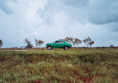 Sandy dirt red road with old vintage car with green plants on sides and grey cloudy sky on background in cuba