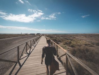 Rear view of woman walking on boardwalk against blue sky