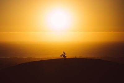Silhouette man riding motorcycle on mountain against orange sky