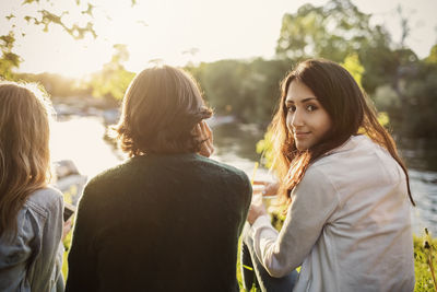 Rear view portrait of female teenager sitting with friends at lakeshore