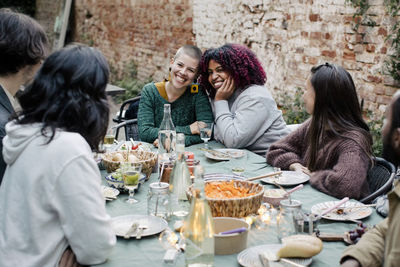 Friends talking to multiracial women sitting at dining table during dinner party in back yard