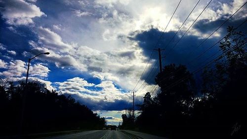 Electricity pylons on road against cloudy sky