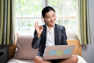 Young woman using mobile phone while sitting on window