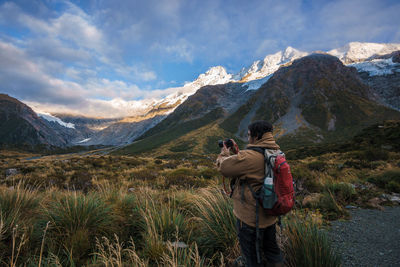 Side view of backpacker photographing field by mountains against sky