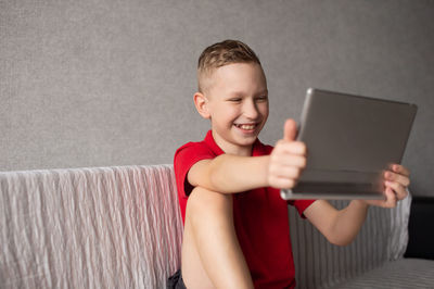 A happy boy in a red t-shirt is sitting on the couch with a tablet and laughing