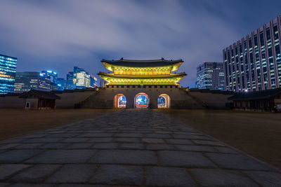 Illuminated gate and buildings against cloudy sky at night