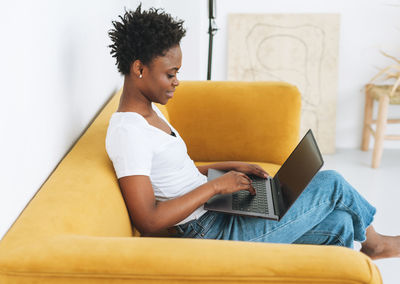 Beautiful african woman in white t-shirt and blue jeans using laptop on yellow sofa in interior