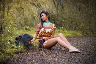 Smiling young woman in traditional clothing sitting by raccoon in forest