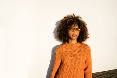 Full length of thoughtful pretty ethnic woman with curly afro hair wearing vivid orange sweater looking away leaning against white modern concrete wall background