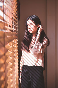 Thoughtful young woman looking through window blinds at home