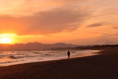 Silhouette person walking at beach against cloudy orange sky during sunset