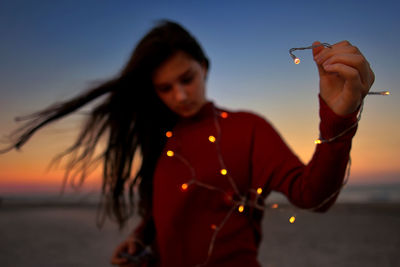 Teenage girl holding illuminated string light while standing at beach during sunset