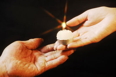 Cropped hand giving lit tea light to person against black background