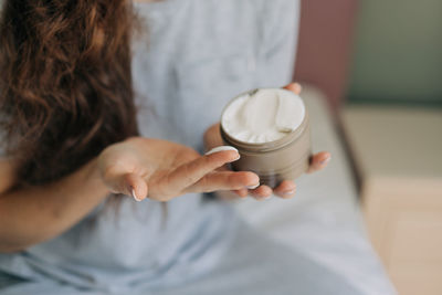 An unrecognizable woman holds a jar of cream and uses cream on her hands.